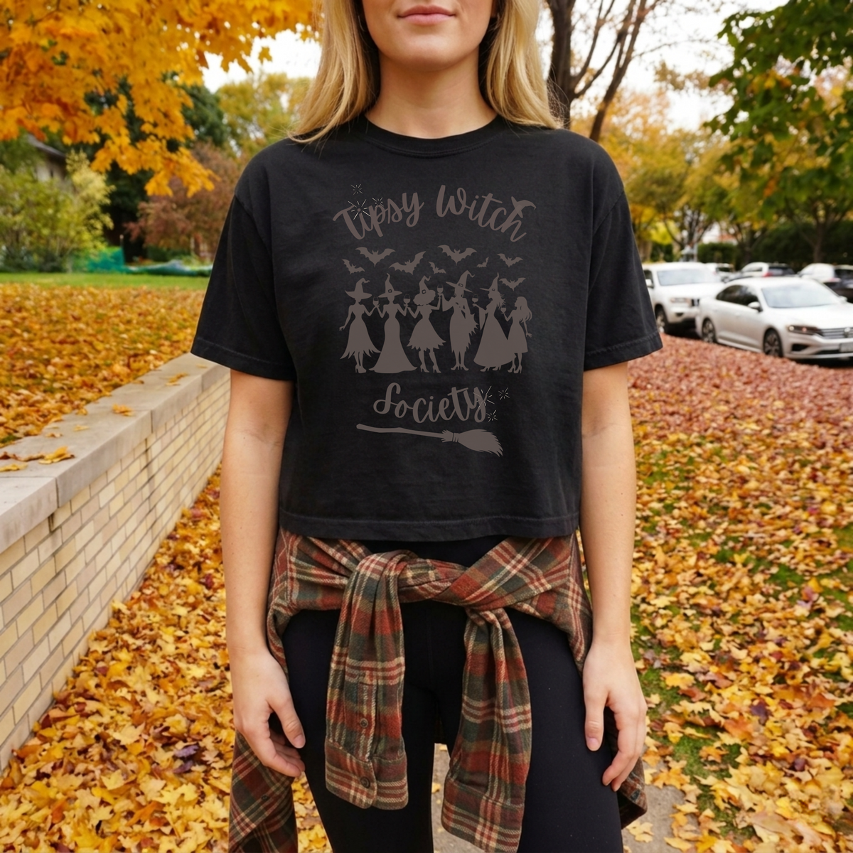 A woman wearing a black graphic t-shirt with a distressed gray print that reads 'Tipsy Witch Society' with silhouettes of witches and bats, styled with a plaid shirt tied around her waist, set against an autumn street background covered in fallen leaves.