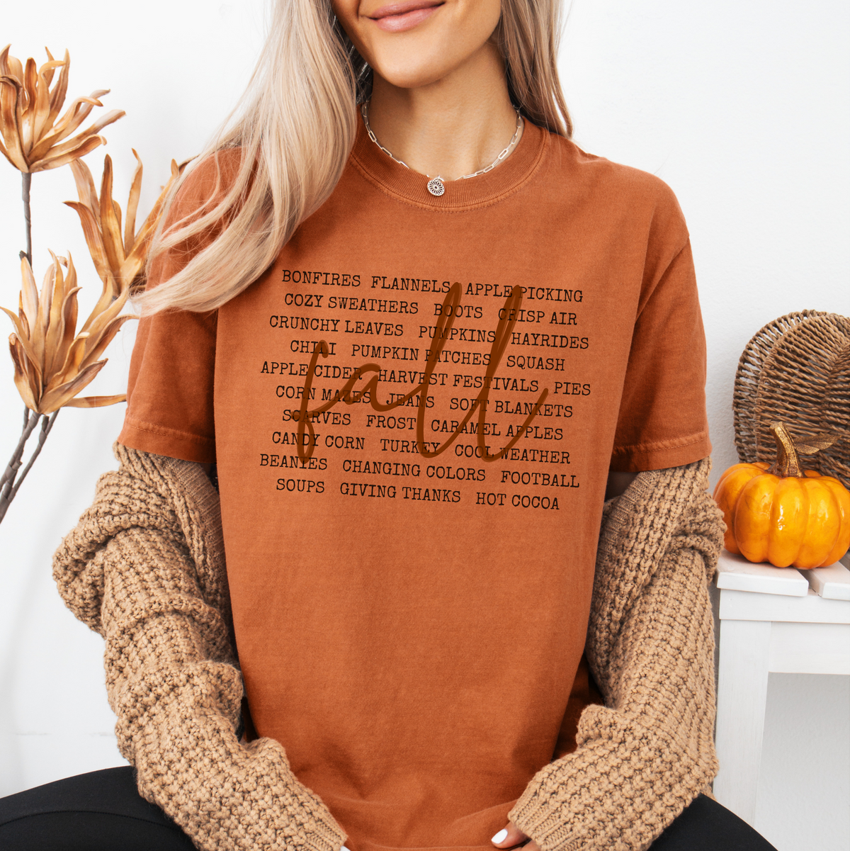 A woman wearing a rust-orange graphic t-shirt with a black text list on the front, layered under a chunky beige knit sweater. She is posed in a cozy, autumn-themed setting with dried flowers and a small pumpkin.