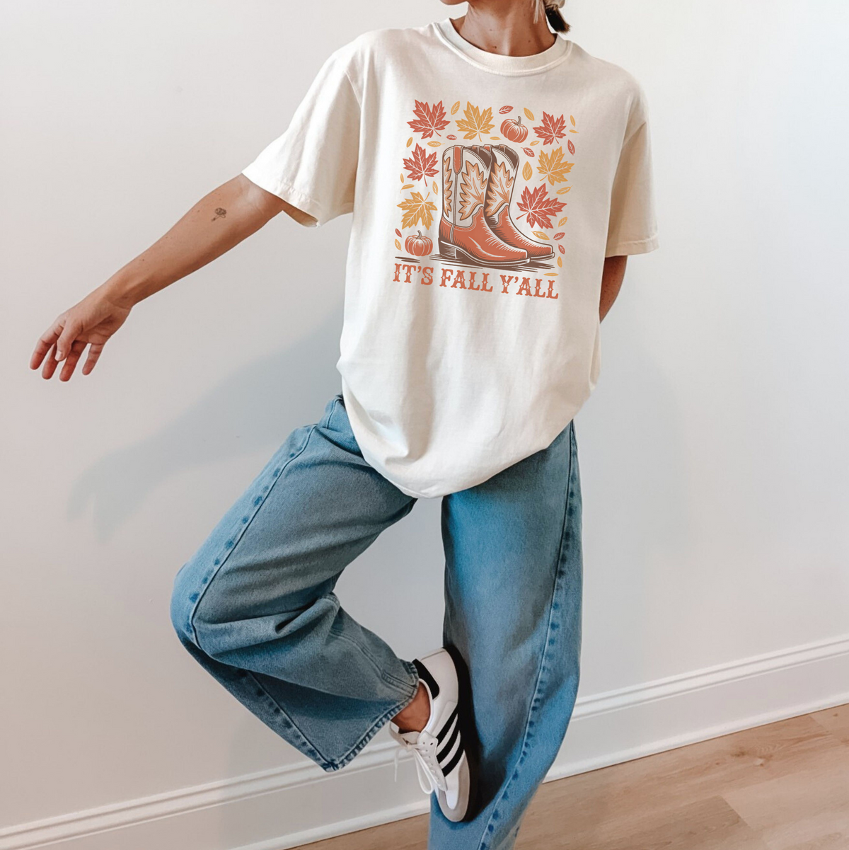 A person modeling a cream-colored graphic t-shirt with a fall-themed boot print and the text 'IT'S FALL 'AALL', paired with wide-leg blue jeans and white sneakers, standing against a plain white wall.