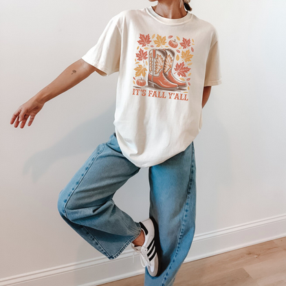 A person modeling a cream-colored graphic t-shirt with a fall-themed boot print and the text 'IT'S FALL 'AALL', paired with wide-leg blue jeans and white sneakers, standing against a plain white wall.