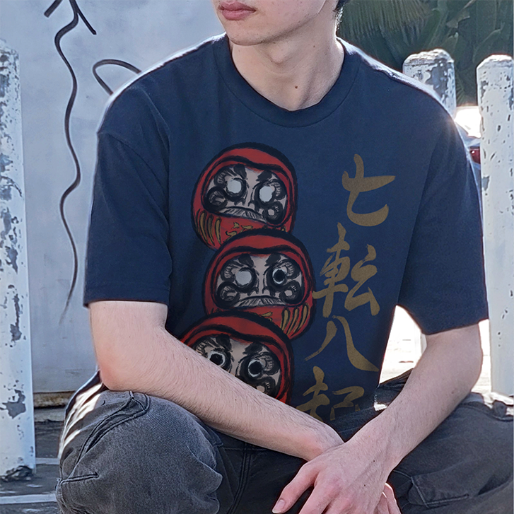 A person crouching outdoors wearing a navy blue graphic T-shirt with three stacked Daruma doll illustrations and gold Japanese characters, paired with dark gray cargo pants and black shoes.