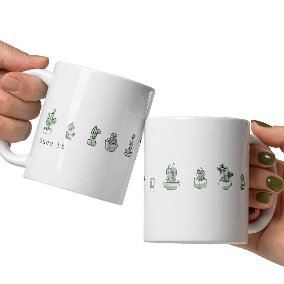 Two white mugs with cactus illustrations held by two hands against a white background