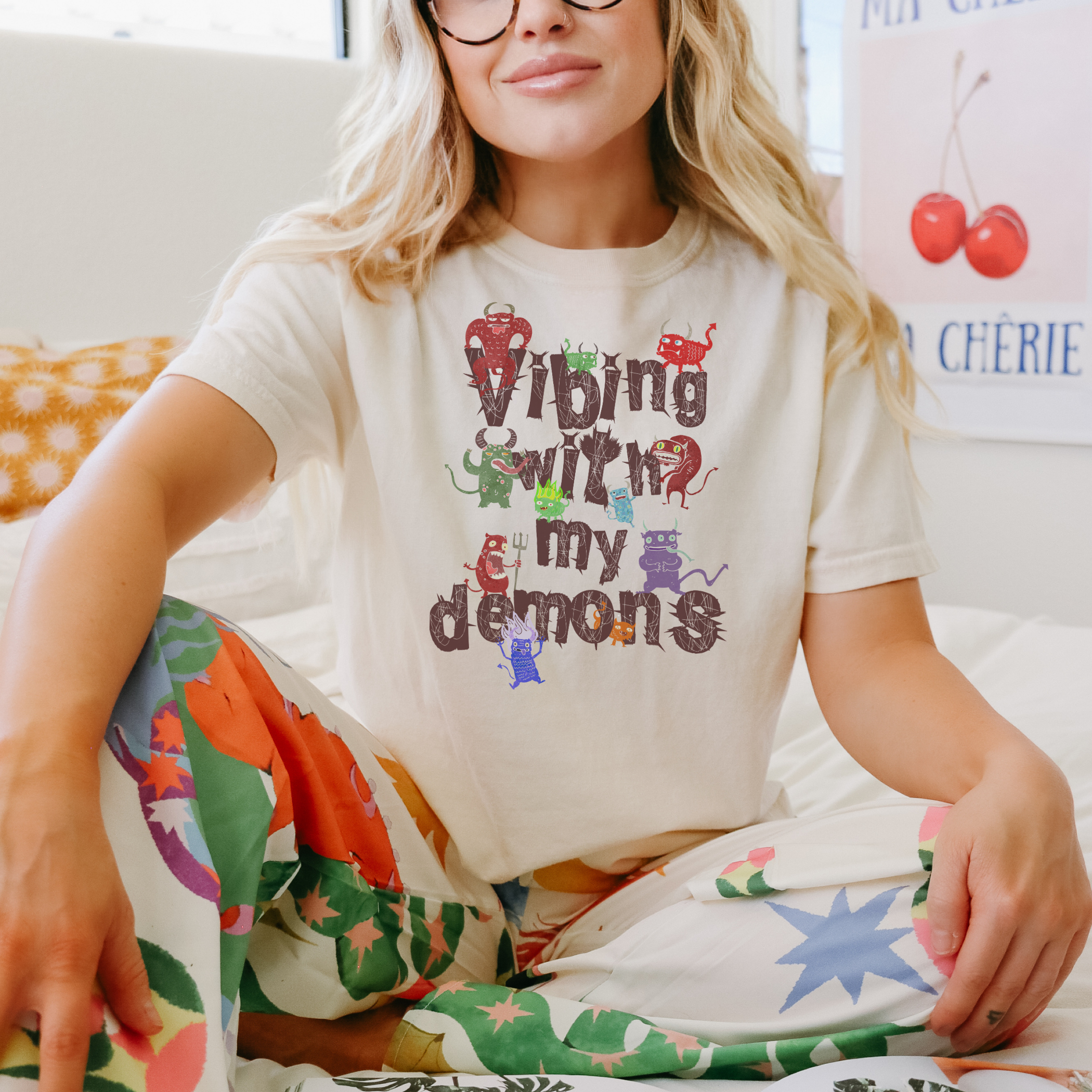 Person wearing a white t-shirt with colorful text "Vibing with my demons" and floral pants sitting on a bed
