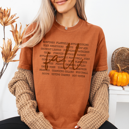 A woman wearing a rust-orange graphic t-shirt with a black text list on the front, layered under a chunky beige knit sweater. She is posed in a cozy, autumn-themed setting with dried flowers and a small pumpkin.