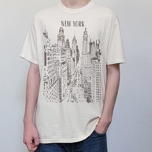 A person wearing a cream-colored short-sleeve t-shirt with a black line art graphic of the New York City skyline and the text 'NEW YORK' printed on the chest, paired with dark blue jeans.