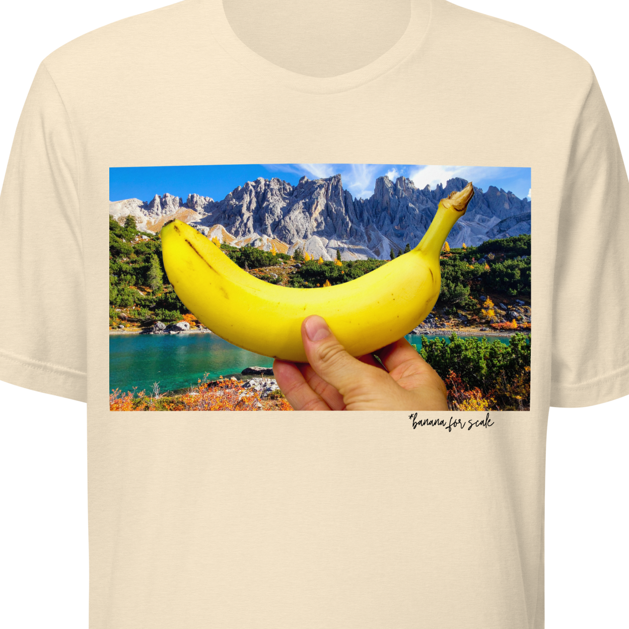 Beige t-shirt with a scenic print of a hand holding a banana over a mountain lake.