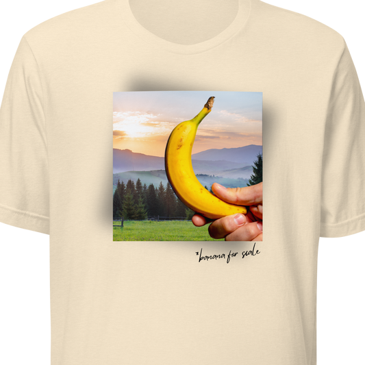 Beige t-shirt with a graphic of a hand holding a banana and a sunset landscape on a white background